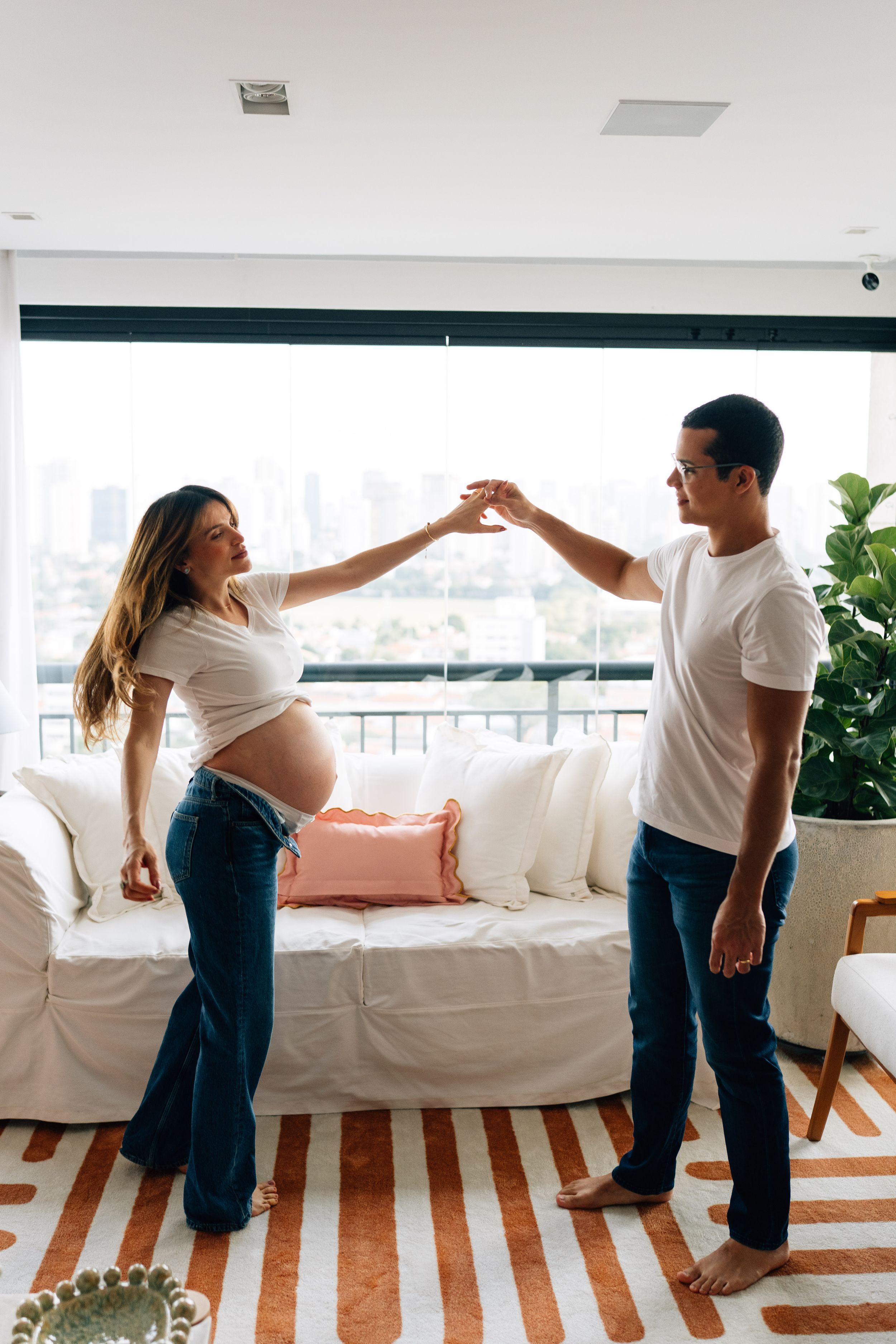 Pregnant woman dancing with partner in modern living room, wearing white shirts and jeans, city view visible through window.