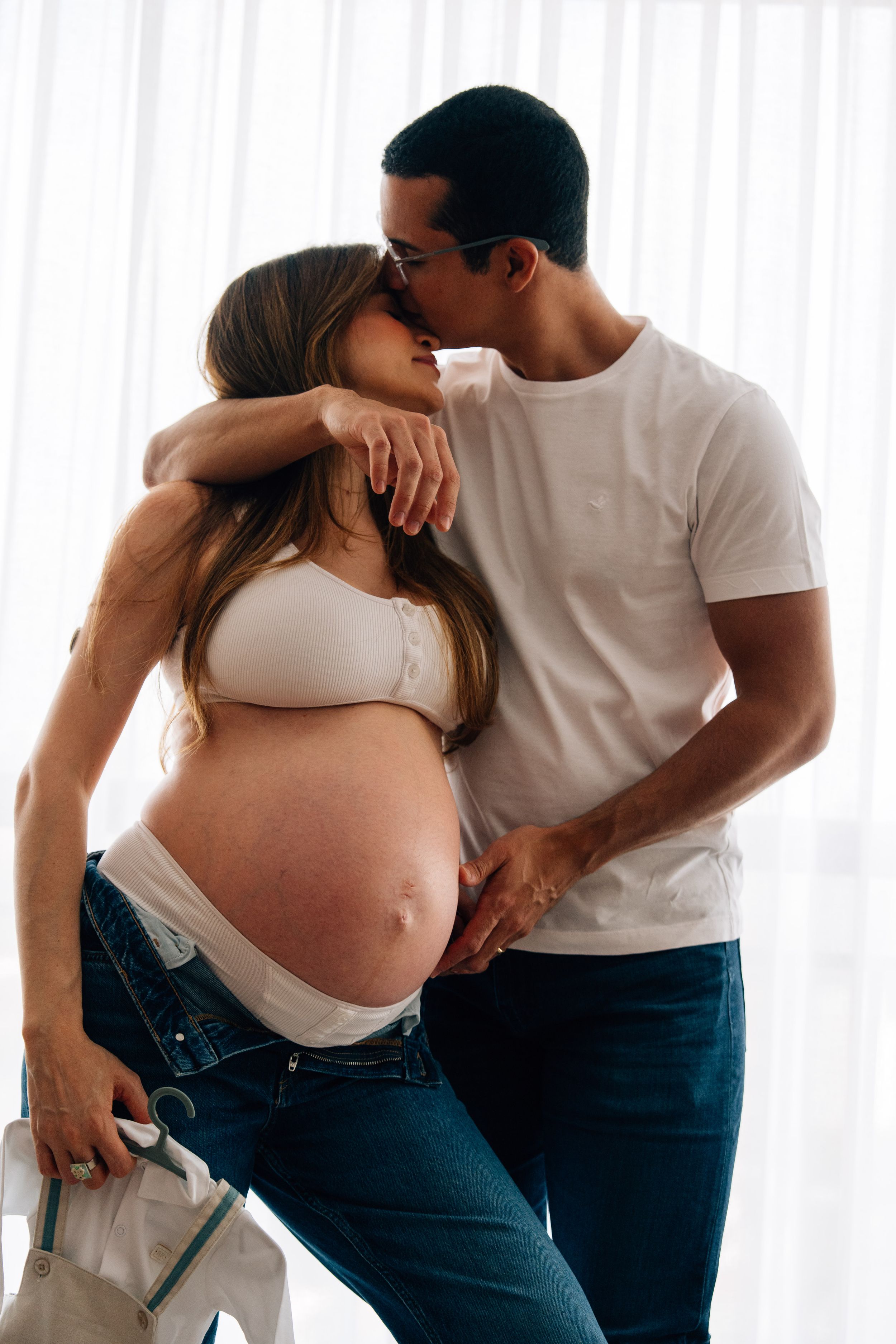 A man tenderly kisses a pregnant woman's head while embracing her baby bump, both wearing white tops and jeans.