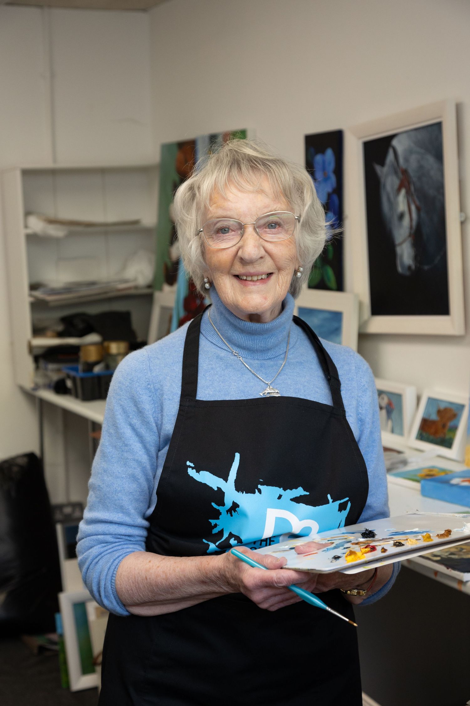 Female artist in her studio, holding a palette in her hands and surrounded by her artwork on the walls.