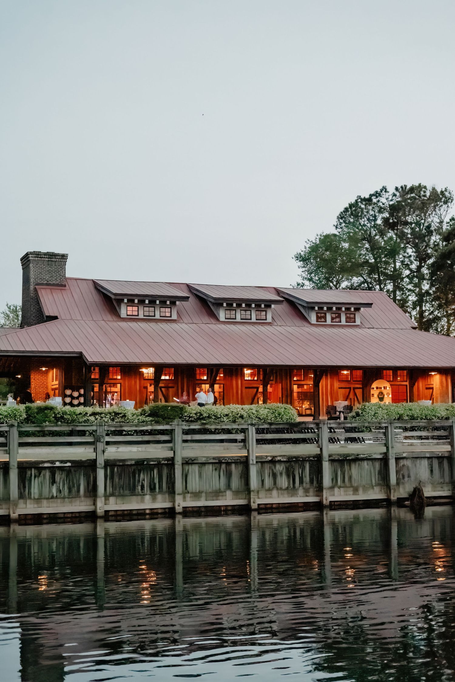 Rustic waterfront lodge with warm glowing windows, red metal roof, and wooden dock reflecting on calm water at dusk.