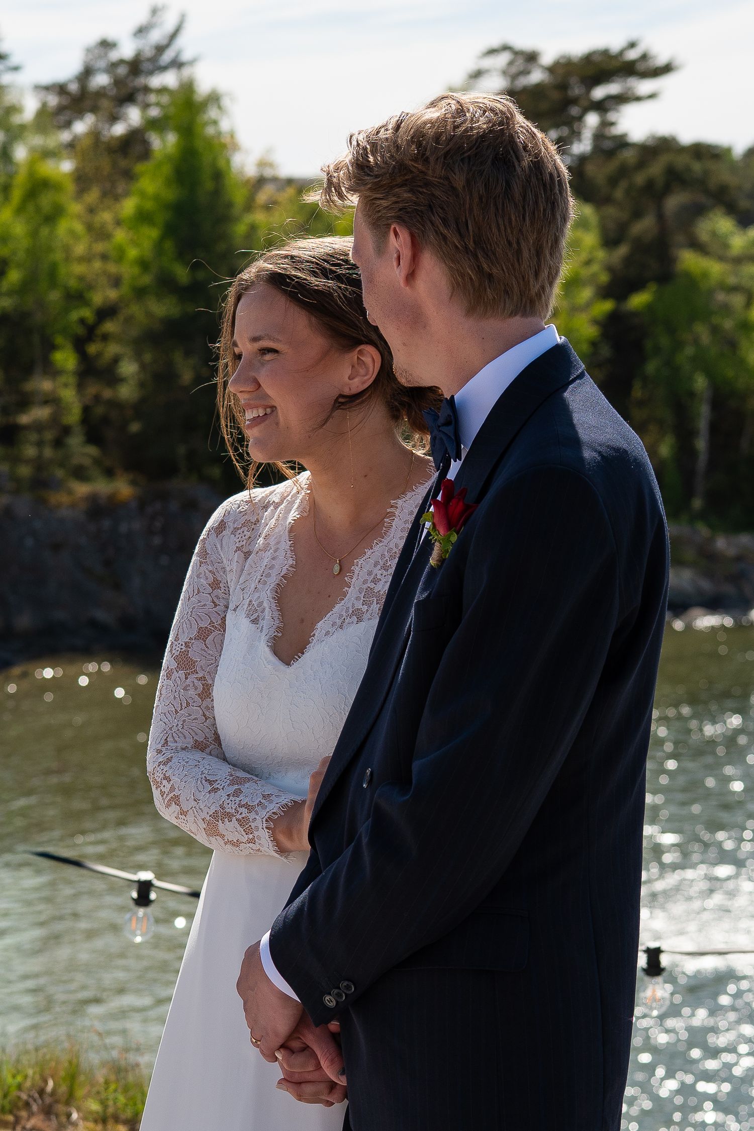 A smiling bride in a lace dress holds hands with a groom in a navy suit outdoors near a sunlit waterway.