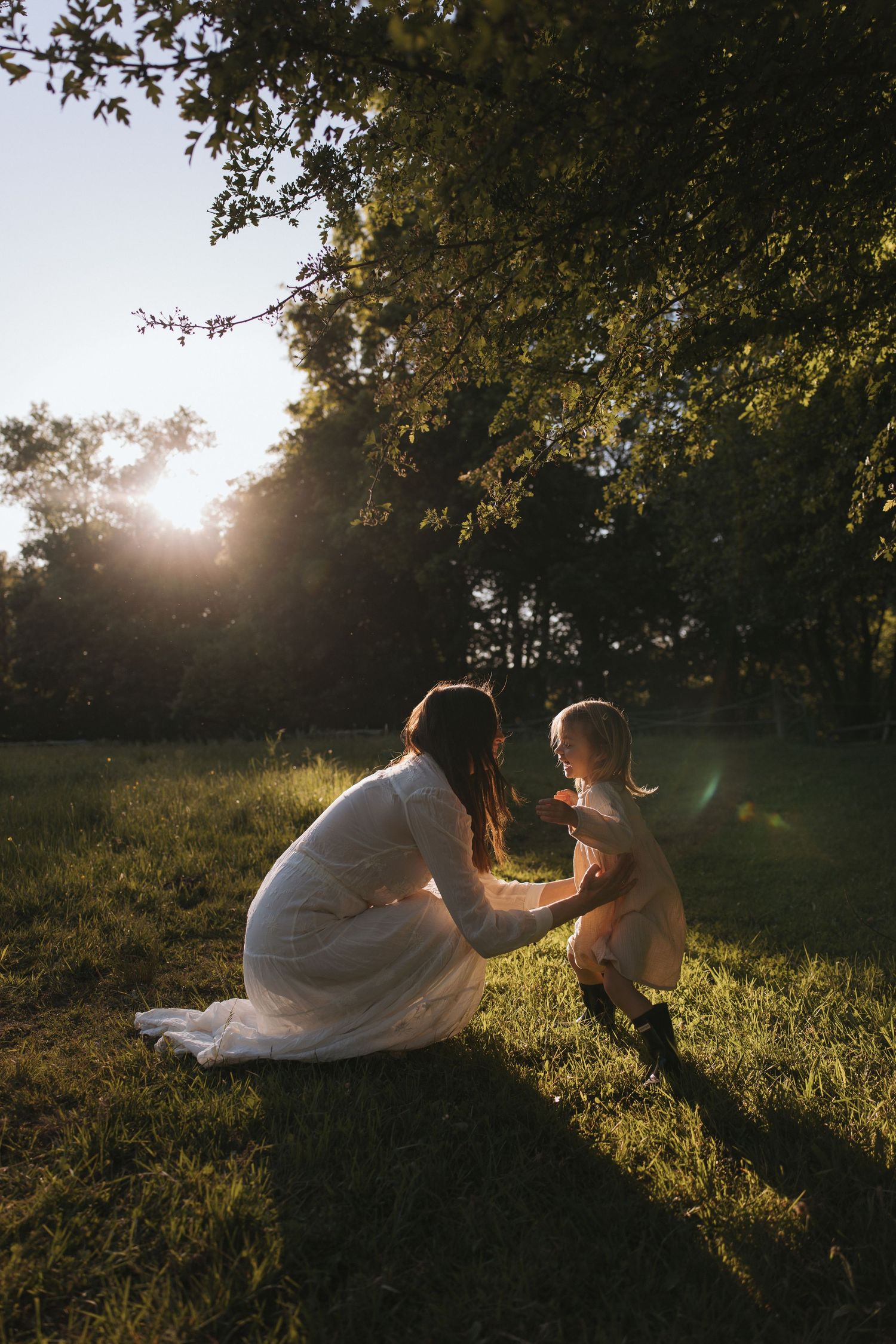 Mother and young daughter share a tender moment in golden sunset light on a lush green lawn beneath tall trees.