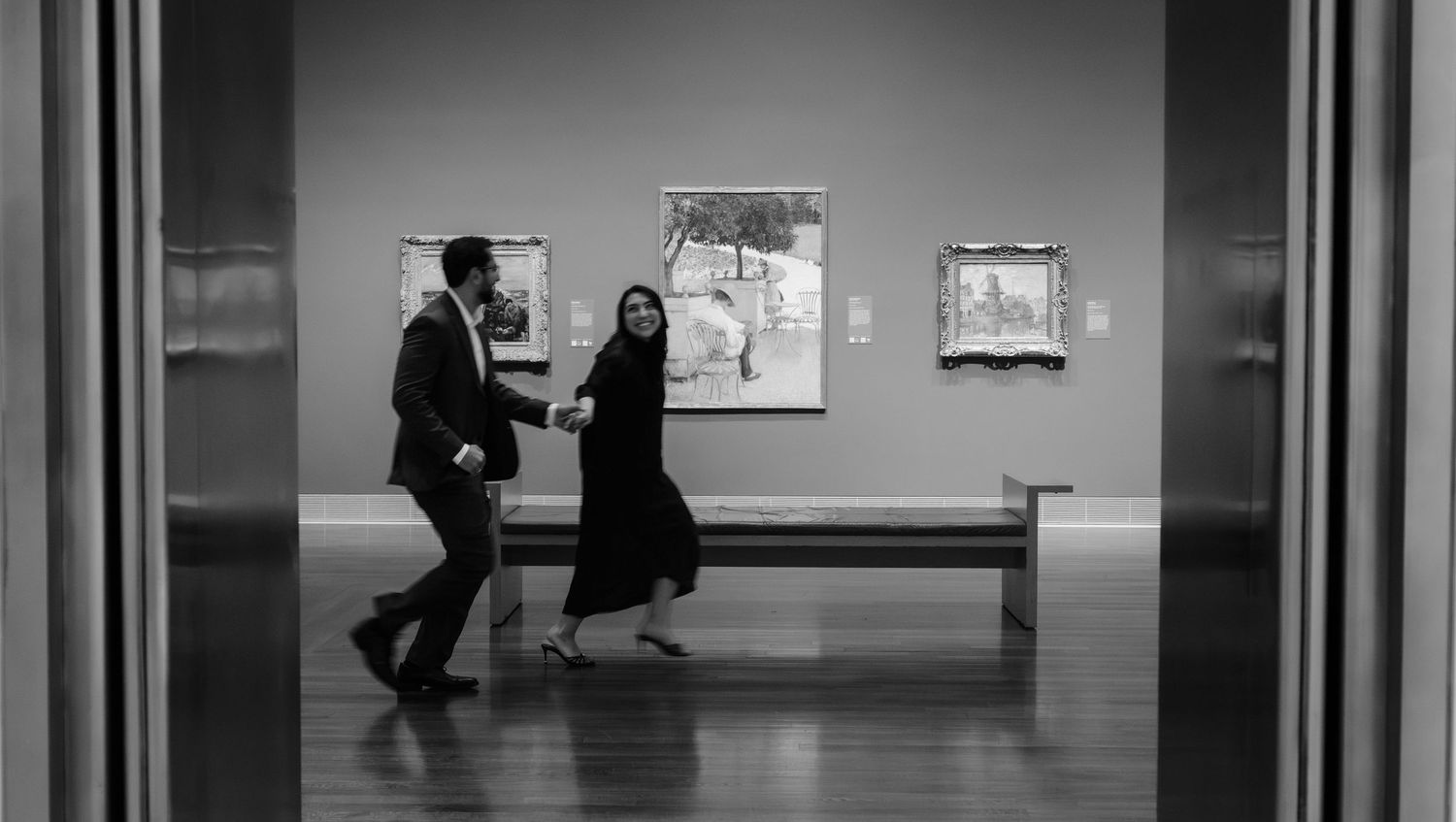 Black and white photo of a man and woman walking through an art museum gallery with paintings on the wall.