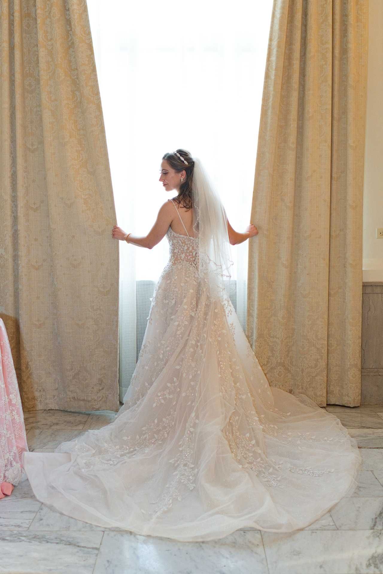 Bride in elegant lace wedding gown with long train and veil stands by curtained window in marble room at The Westin Great Southern Columbus