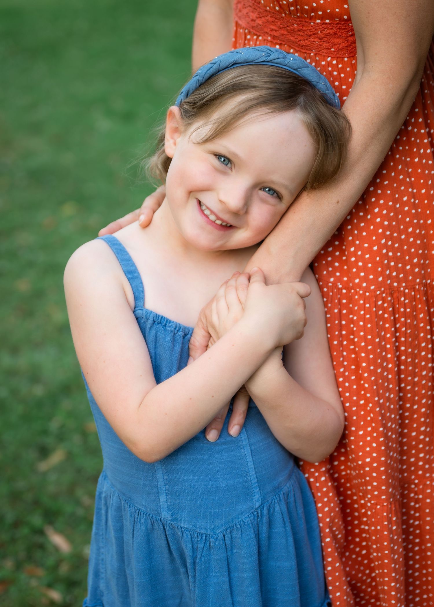 A young girl in a blue dress hugs her mother's arm, smiling warmly outdoors on green grass.