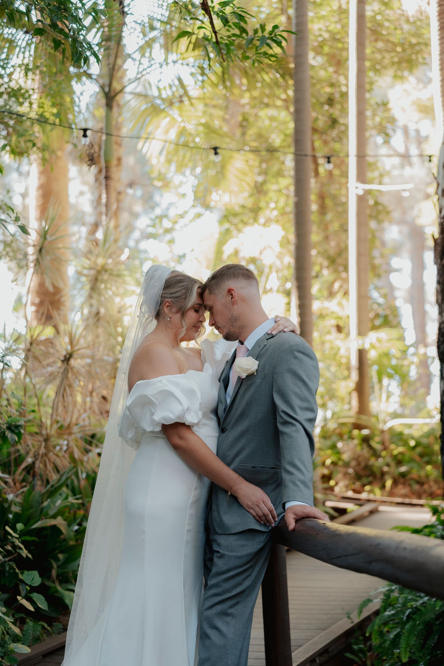 Bride and groom share a romantic forehead touch in a lush tropical garden, bride in off-shoulder gown with veil.