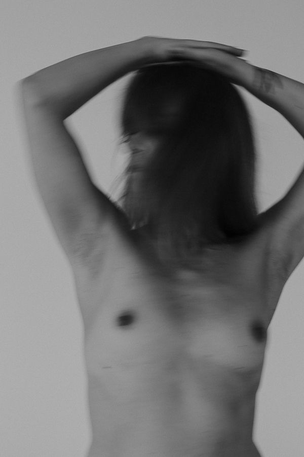 Black and white artistic nude torso photograph with motion blur effect, arms raised above head.