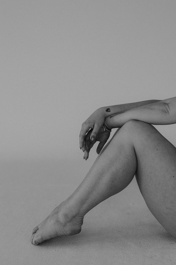 Black and white photo of a person sitting on sand, showcasing bare legs and hands in an artistic pose.