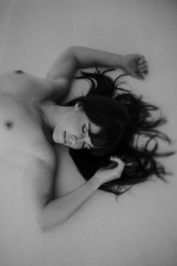 Black and white artistic photo of a person lying down with long dark hair spread out, eyes closed, shirtless.