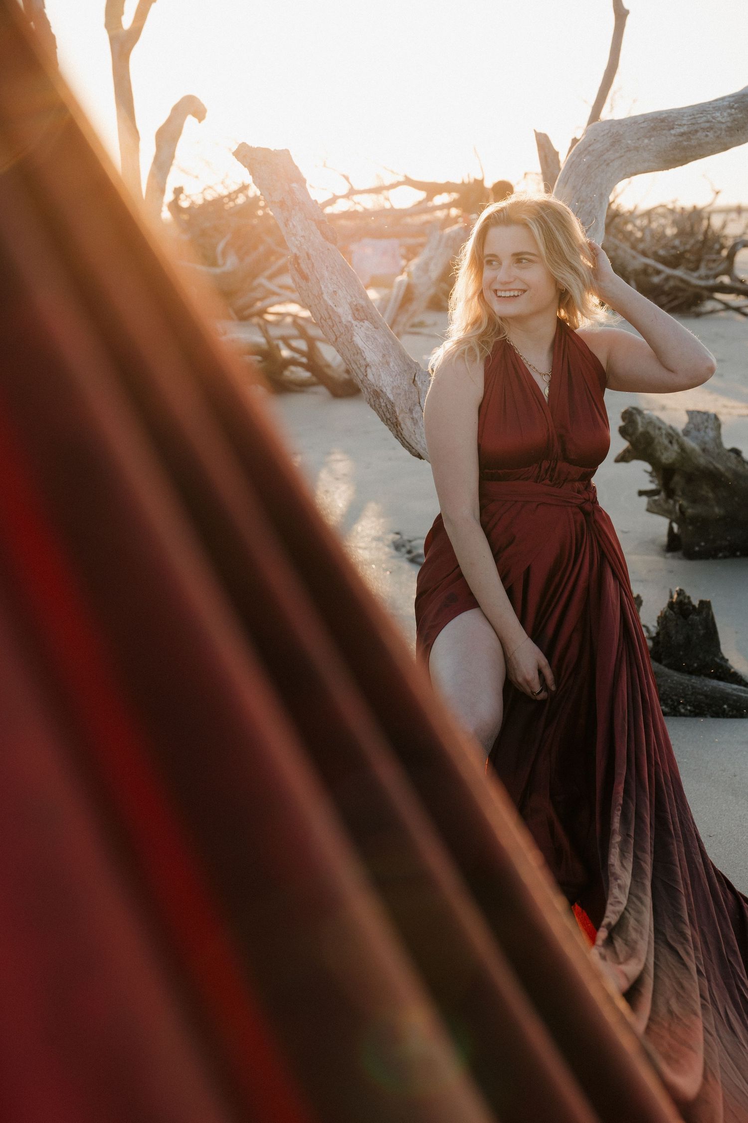 A woman in a red dress with the sun glowing behind her amidst the driftwood