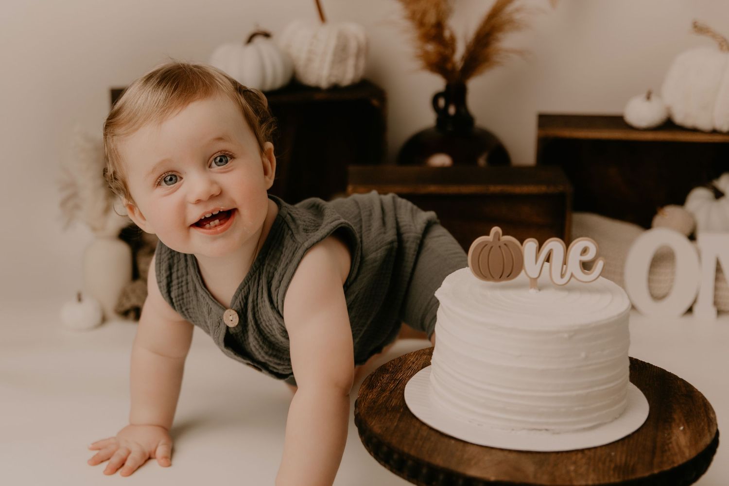 Smiling baby in green romper crawling near a white one cake and pumpkin decorations during a first birthday smash cake session.