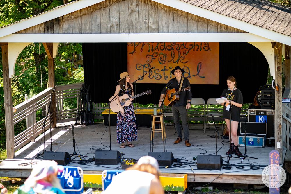 Three musicians perform on an outdoor stage at Mudpond Folk Festival surrounded by lush green trees.