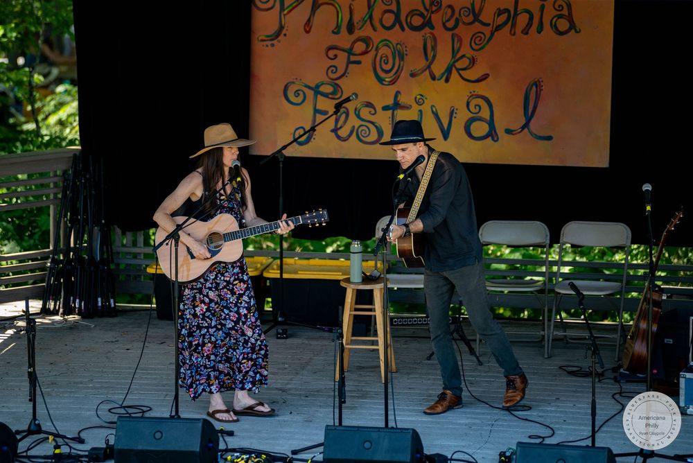 Two musicians perform on stage at the Philadelphia Folk Festival, playing acoustic guitars outdoors.