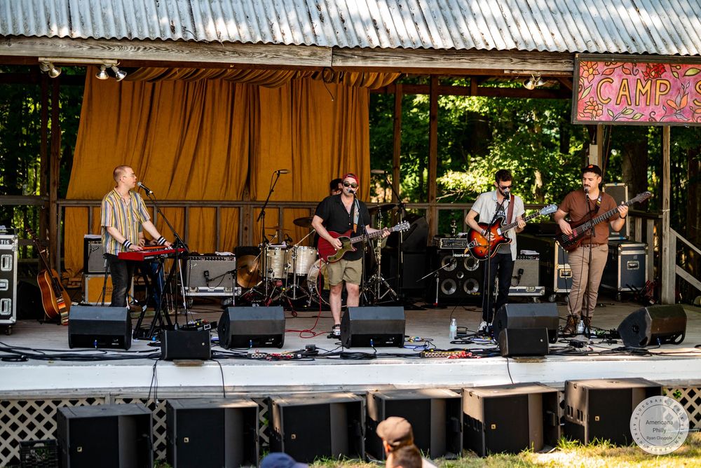 Live band performing on an outdoor stage at a summer music festival surrounded by lush green trees.