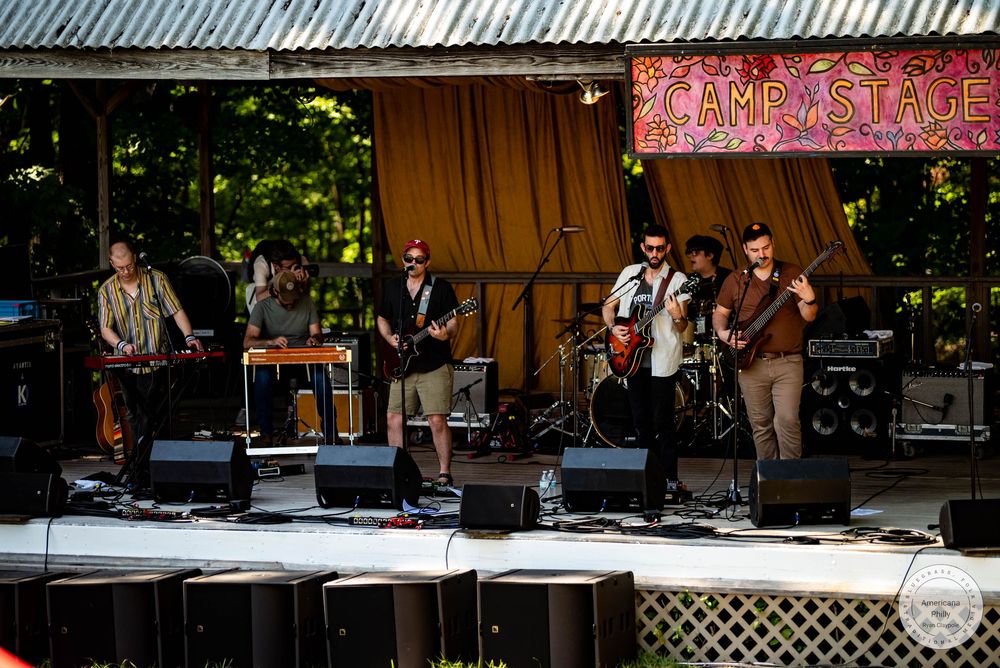 Live band performing on Camp Stage outdoors, with multiple musicians playing guitars and instruments under a shaded pavilion.