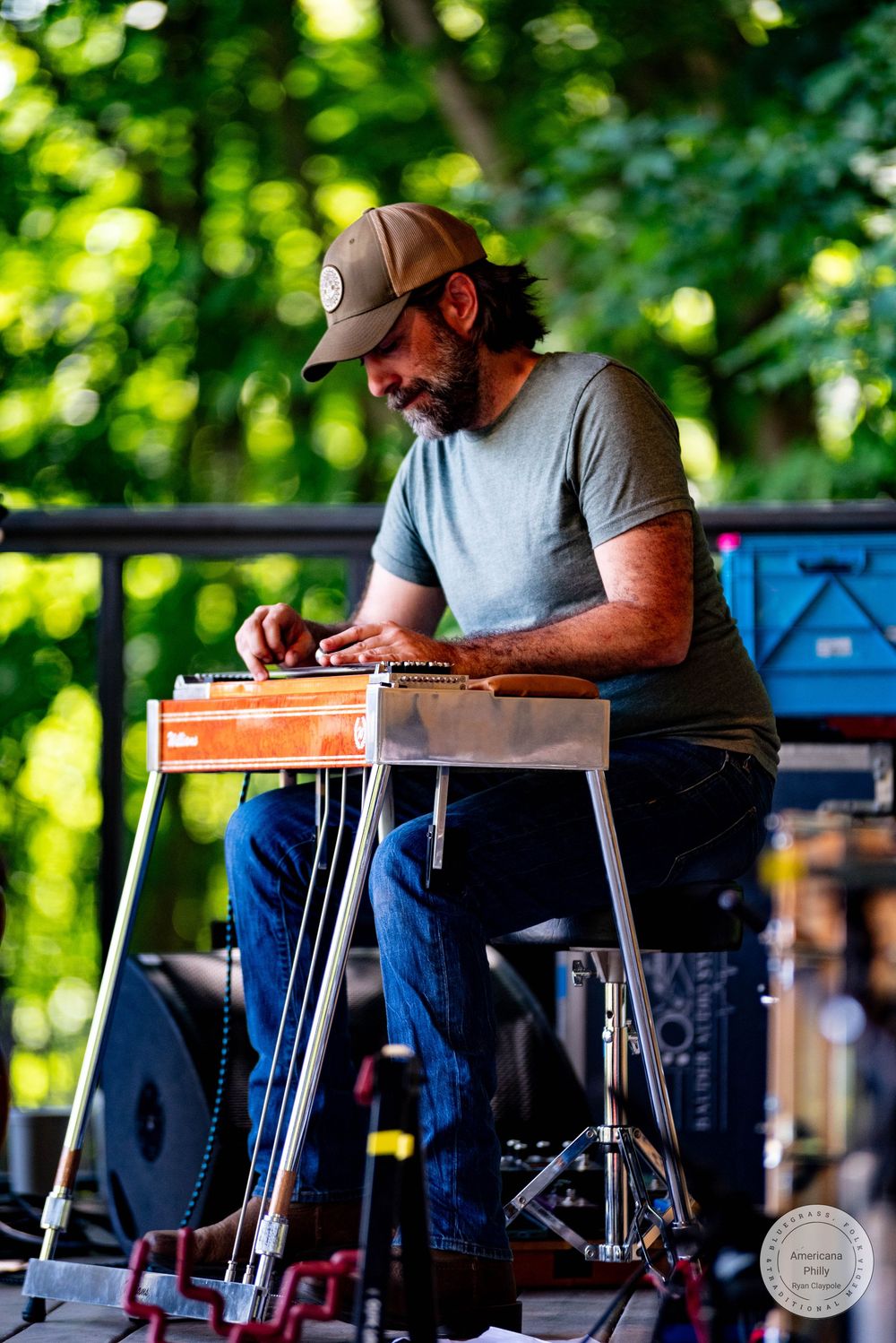 Man playing pedal steel guitar outdoors on stage, wearing a cap and jeans, surrounded by lush green trees.