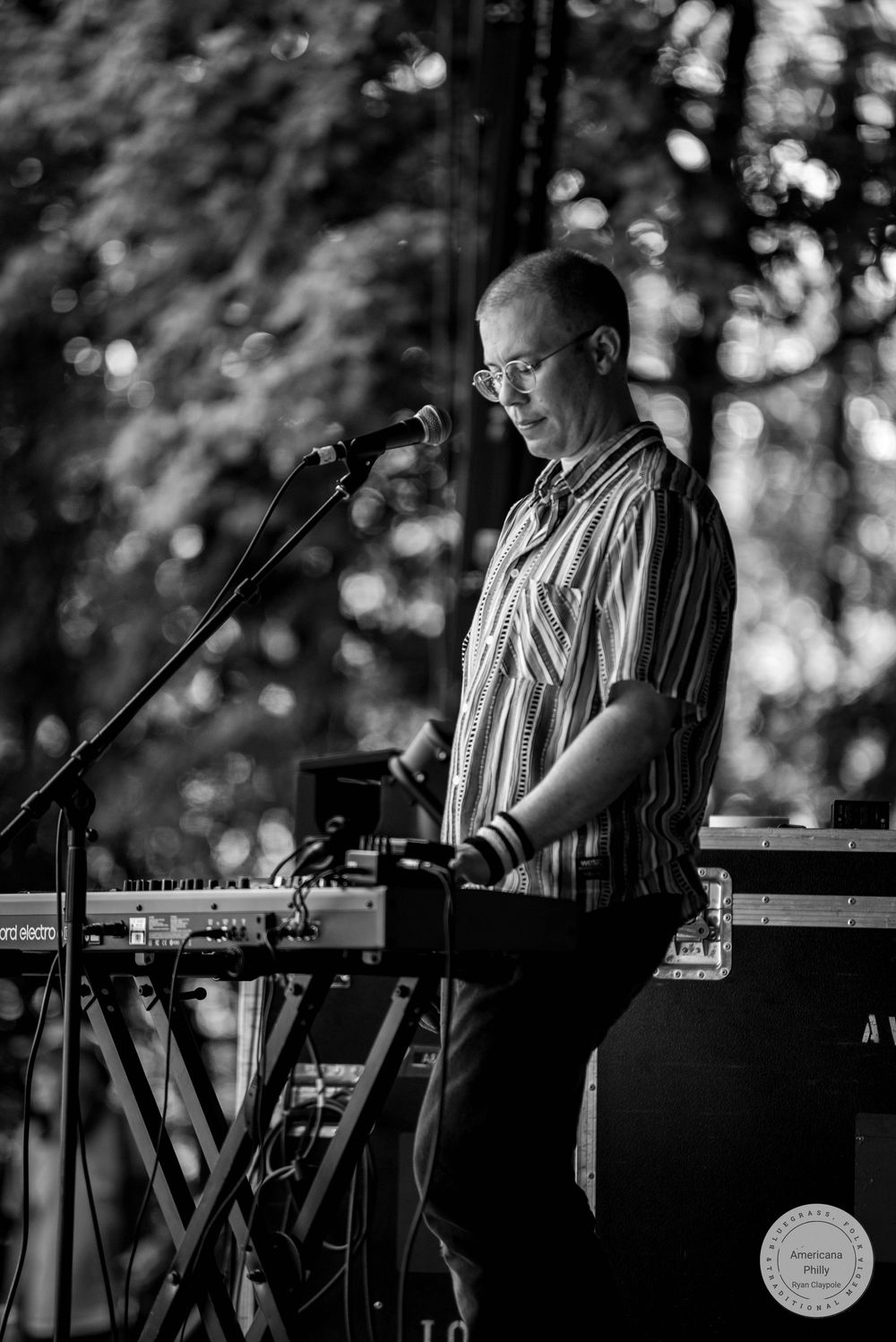 Black and white photo of a bald man with glasses playing keyboard at an outdoor music event.