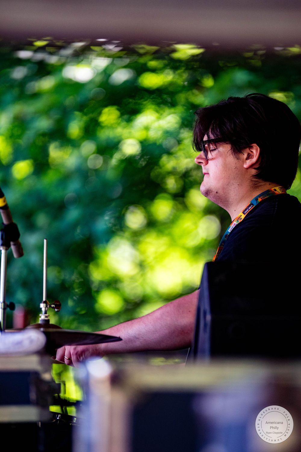 Young drummer with glasses plays cymbals on outdoor stage surrounded by lush green trees.