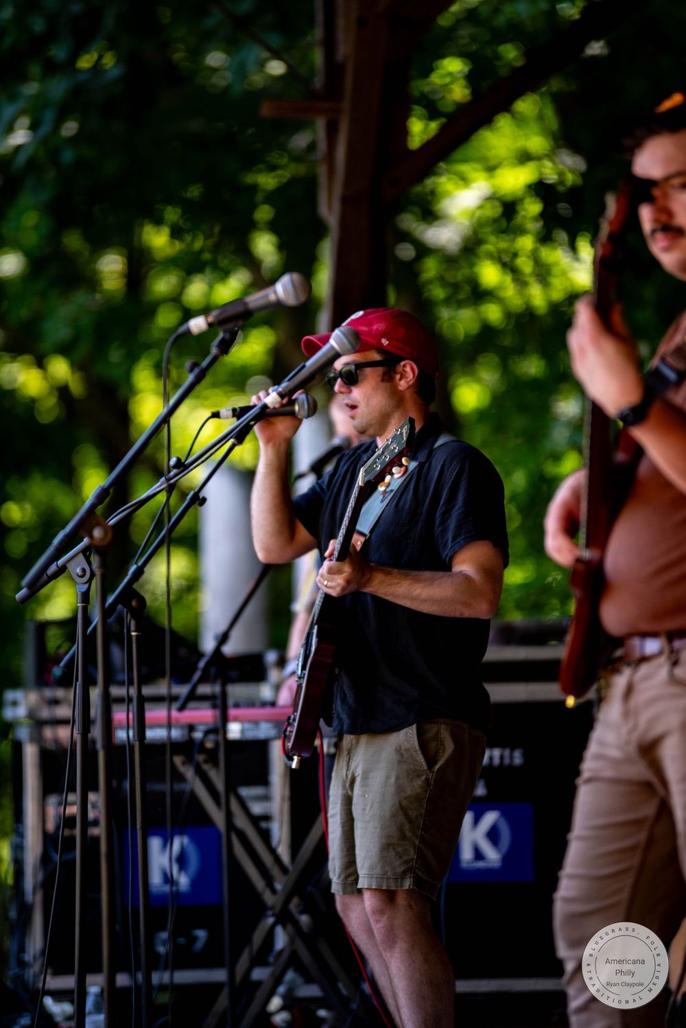 Musician in red cap and sunglasses singing into microphone while playing guitar on outdoor stage.