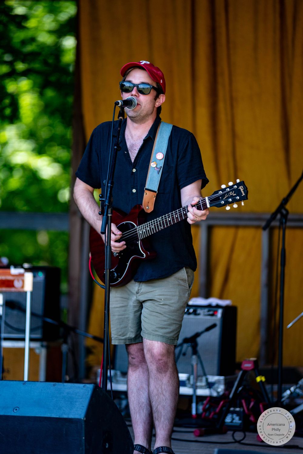 Man playing guitar and singing into microphone on outdoor stage, wearing red cap, sunglasses, and khaki shorts at music festival.