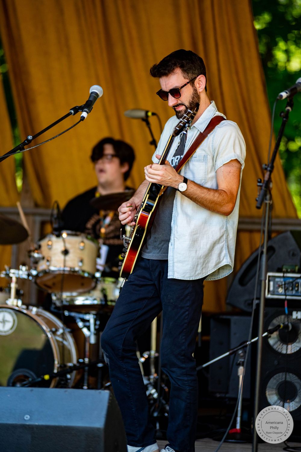 Bearded guitarist in sunglasses plays acoustic guitar on outdoor stage with drummer performing in background.