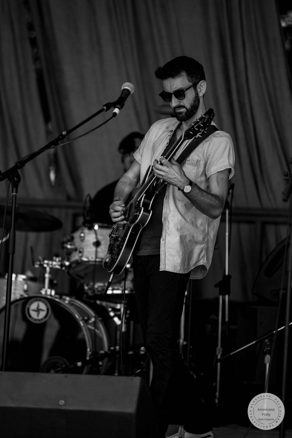 Black and white photo of a bearded guitarist wearing sunglasses performing on stage with a drum kit visible behind him.