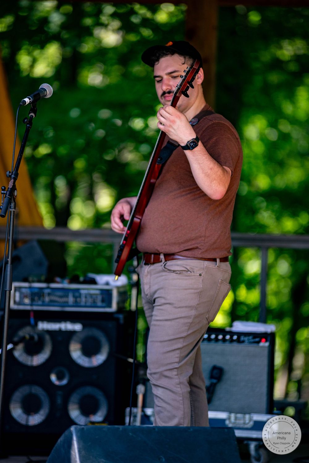 Male guitarist performing on outdoor stage, wearing brown t-shirt and khaki pants, with green trees in background.