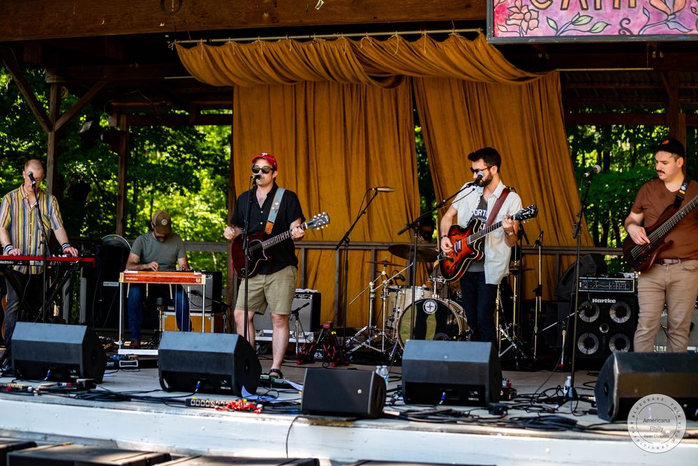 Live band performing on an outdoor stage at a summer music festival, with guitarists and keyboardist under a pavilion.