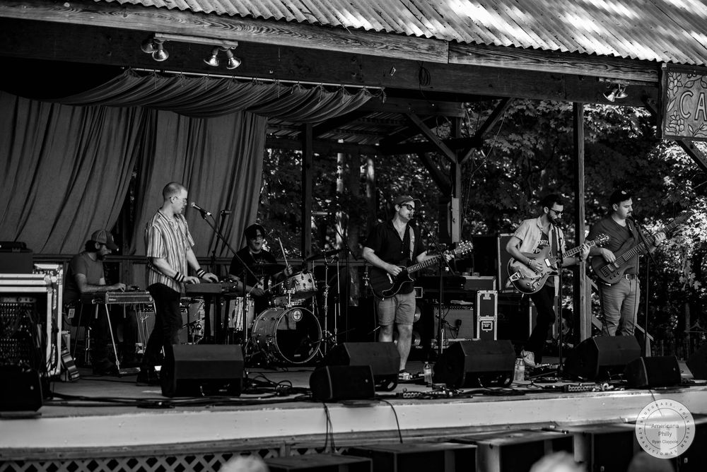 Black and white photo of a live band performing on an outdoor stage surrounded by trees at a music festival.