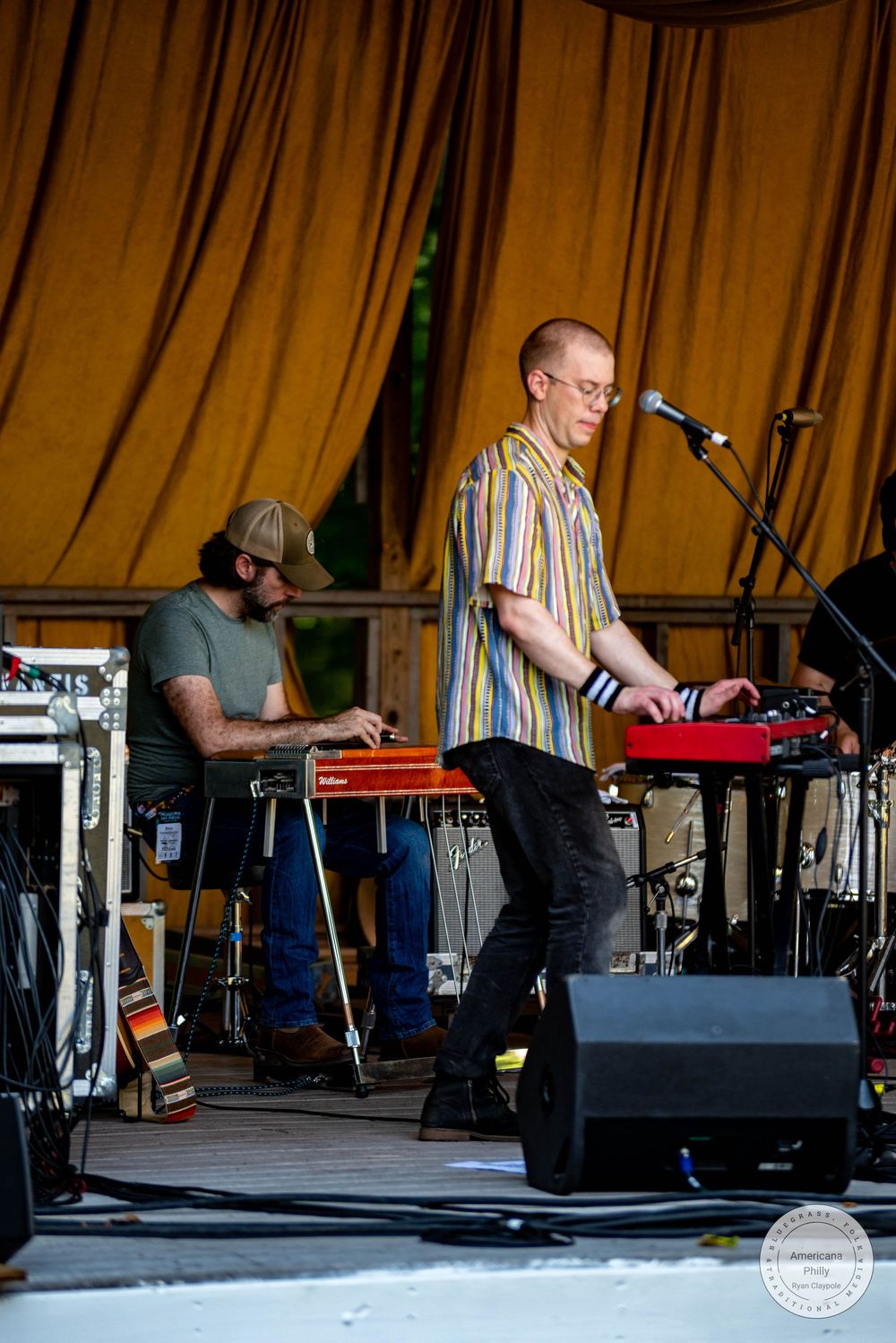 Two musicians perform on stage, one playing a red keyboard and another on a steel guitar, under a yellow draped tent.