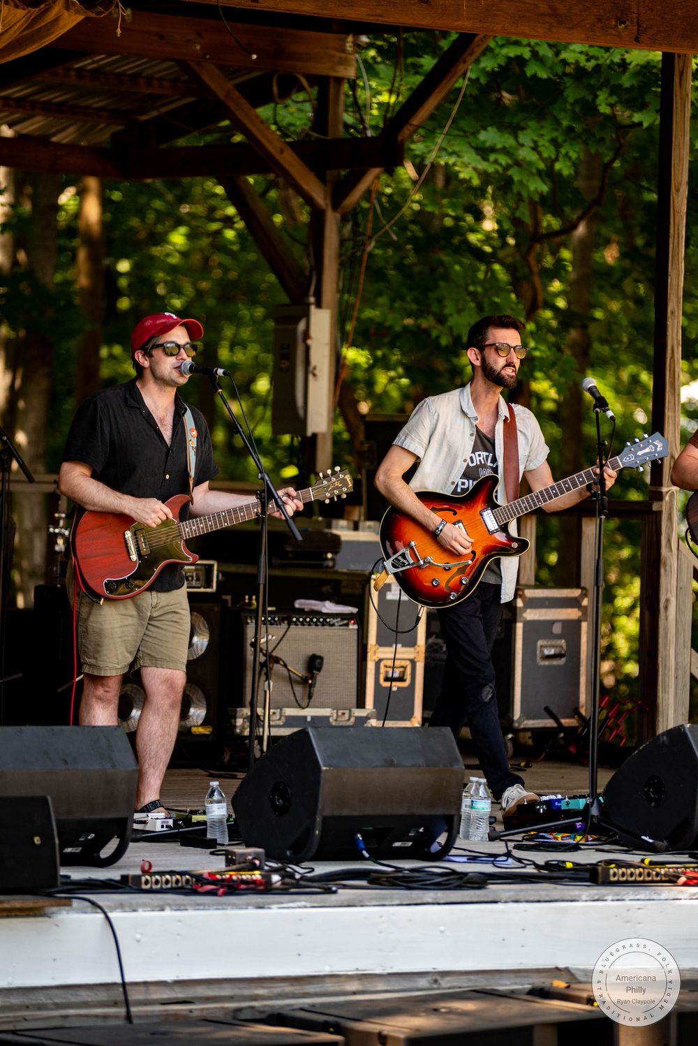 Two guitarists performing on an outdoor stage at a summer music festival surrounded by lush green trees.