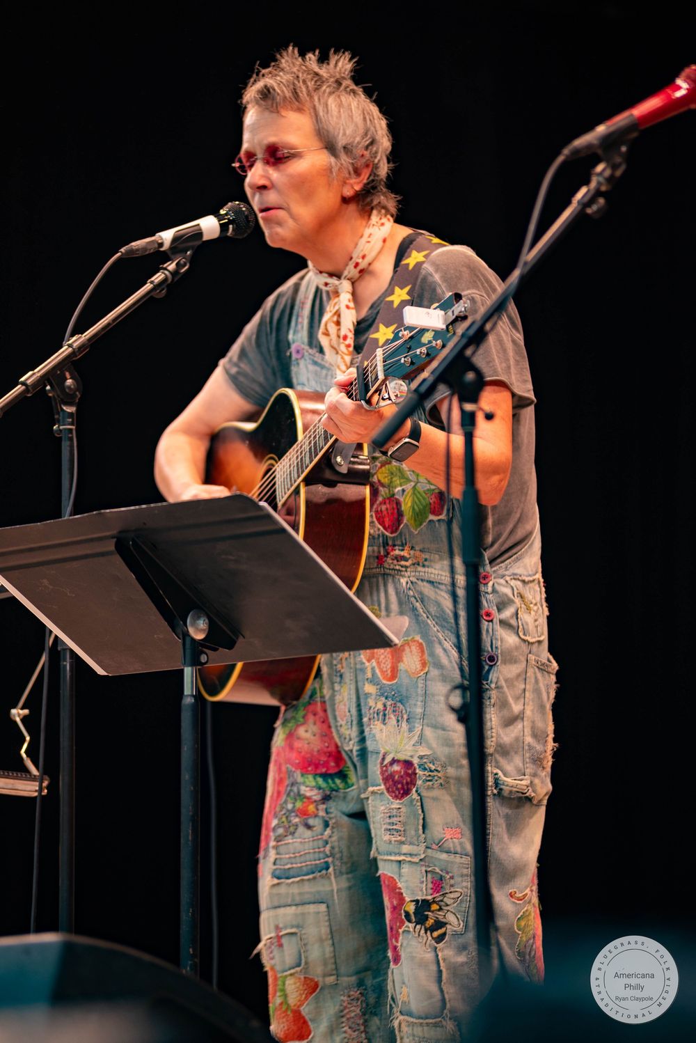 Musician performing acoustic guitar and singing into microphone on stage wearing colorful floral overalls.