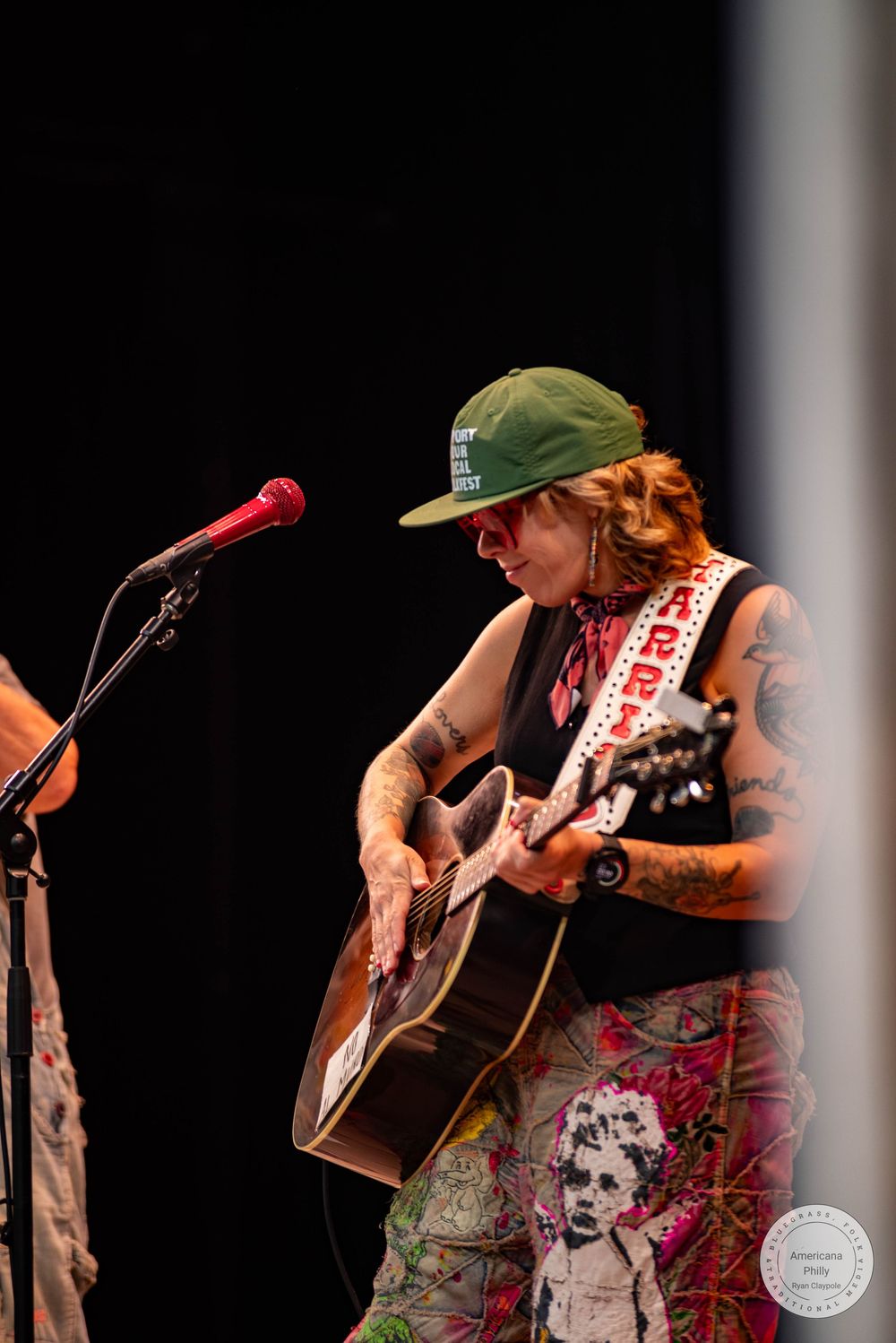 Tattooed guitarist in green cap and printed pants performing on stage with acoustic guitar.