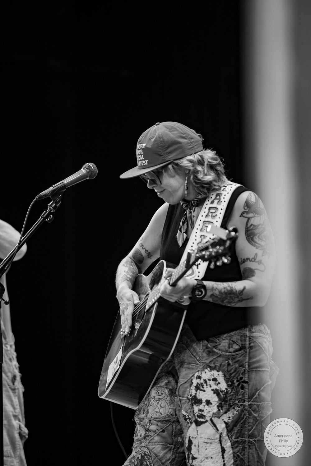Tattooed musician playing acoustic guitar on stage in black and white, wearing a cap and patterned pants.