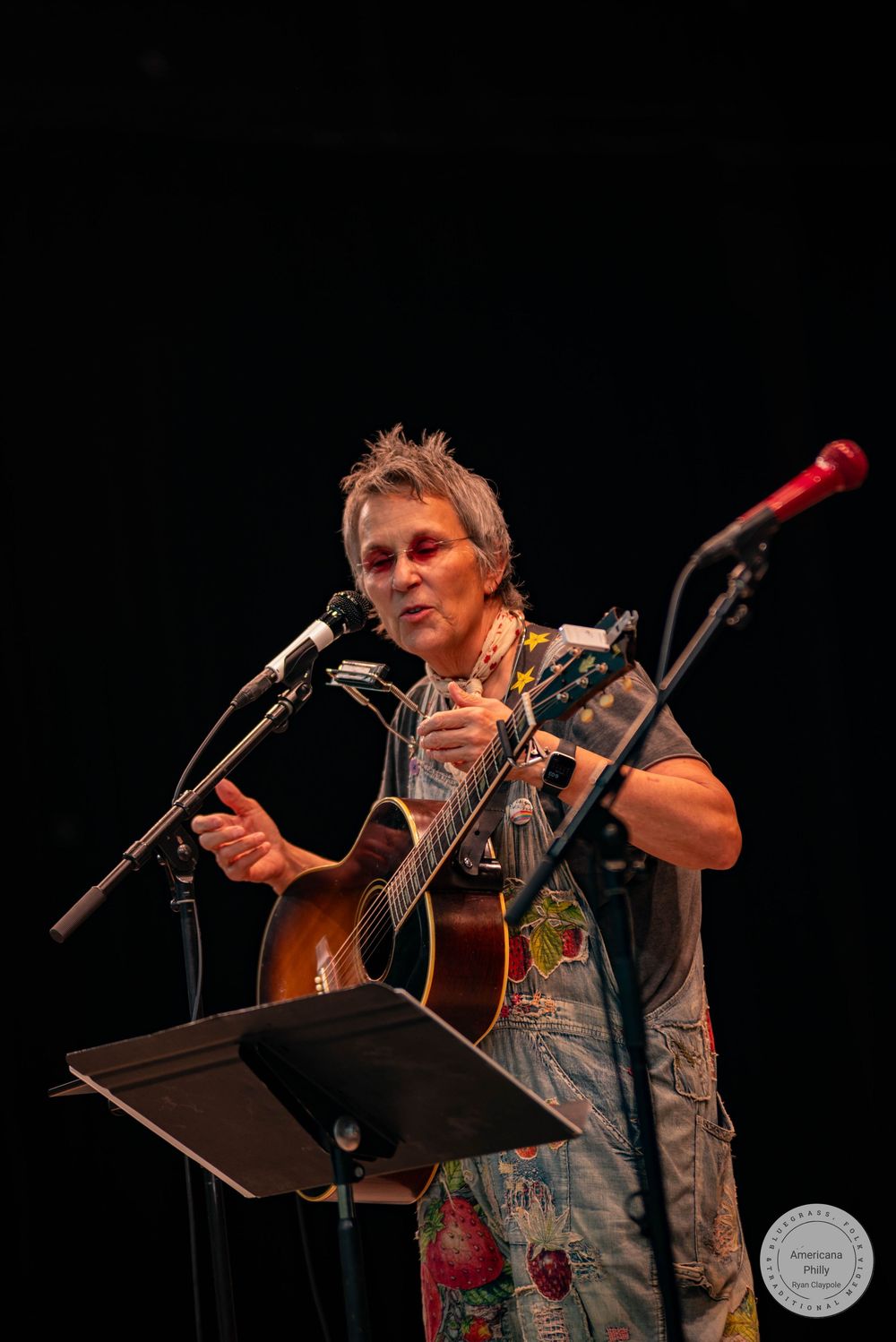 Musician performing on stage with acoustic guitar and microphone, wearing colorful floral shirt under dark concert lighting.