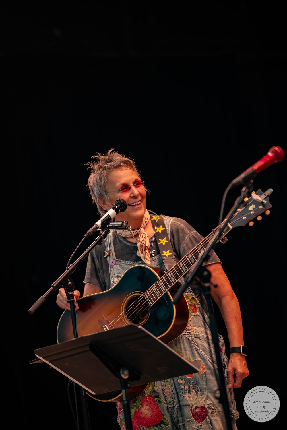 A smiling woman plays acoustic guitar on stage at a microphone stand under warm stage lighting.