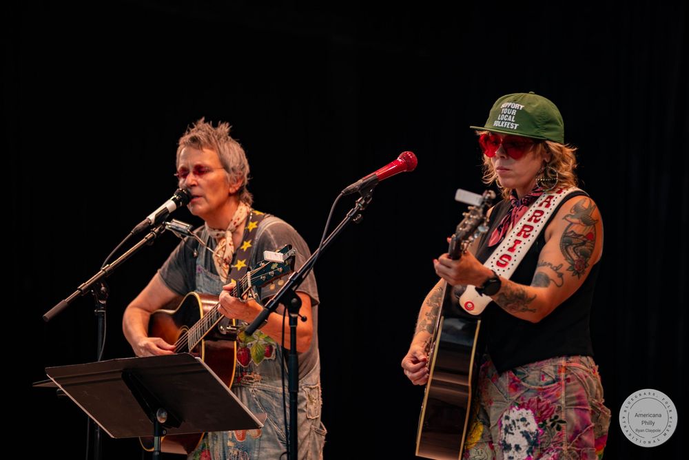 Two musicians playing acoustic guitars and singing into microphones on a dark stage during a live performance.