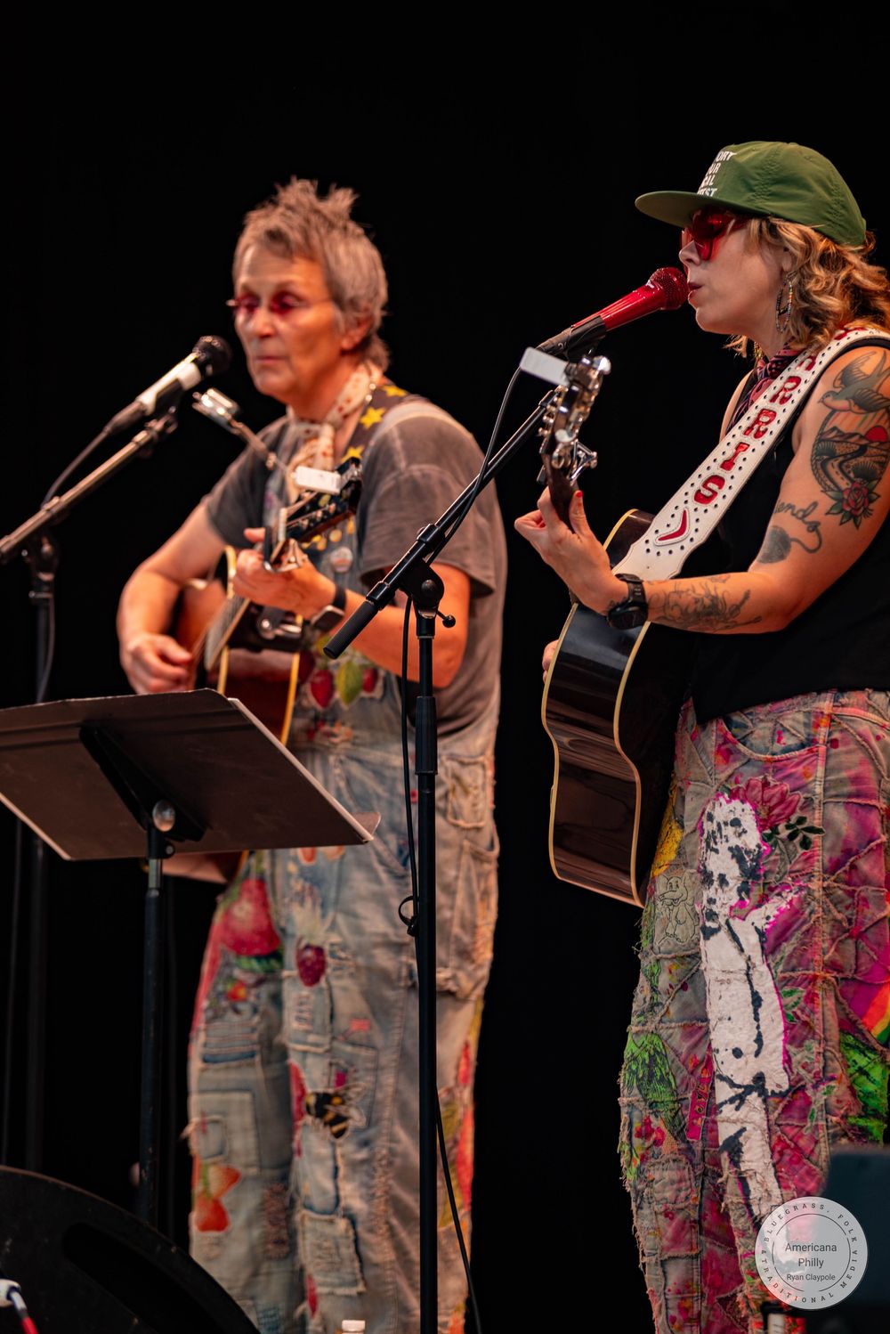 Two guitarists performing on stage, one in floral overalls, one in printed skirt and green cap.