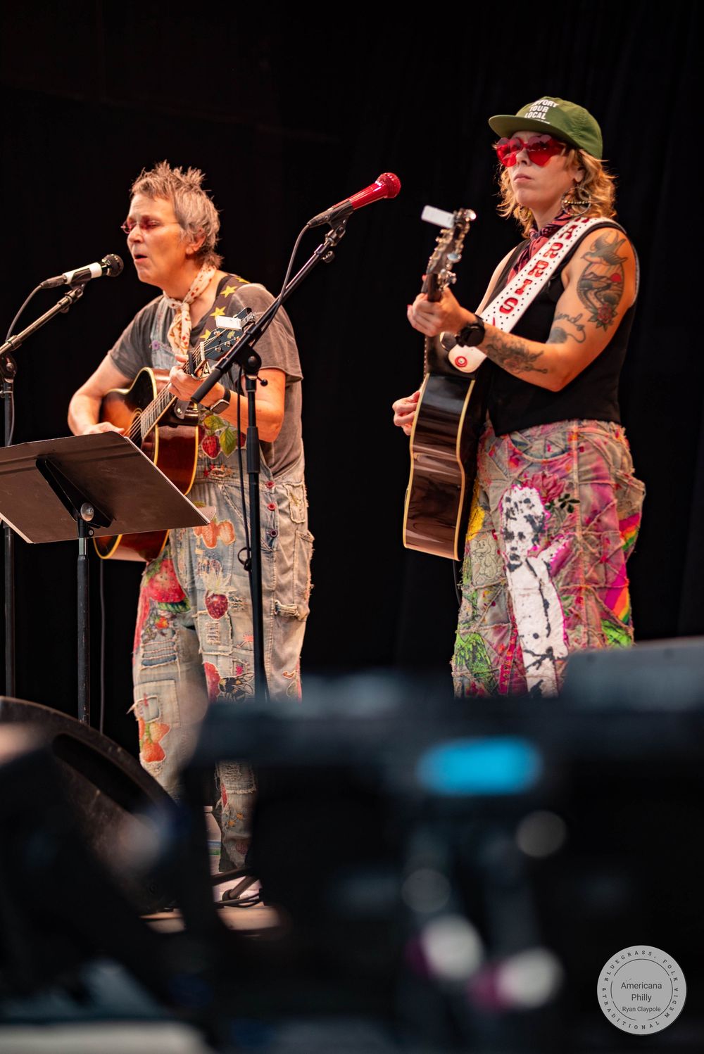 Two musicians perform acoustic guitars on stage, one tattooed in colorful outfit, both at microphones during live concert.