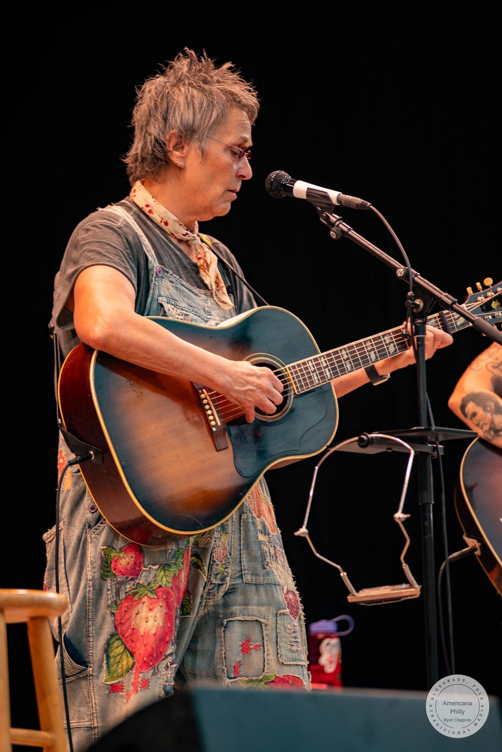 Musician playing blue acoustic guitar on stage wearing floral overalls at a live performance.