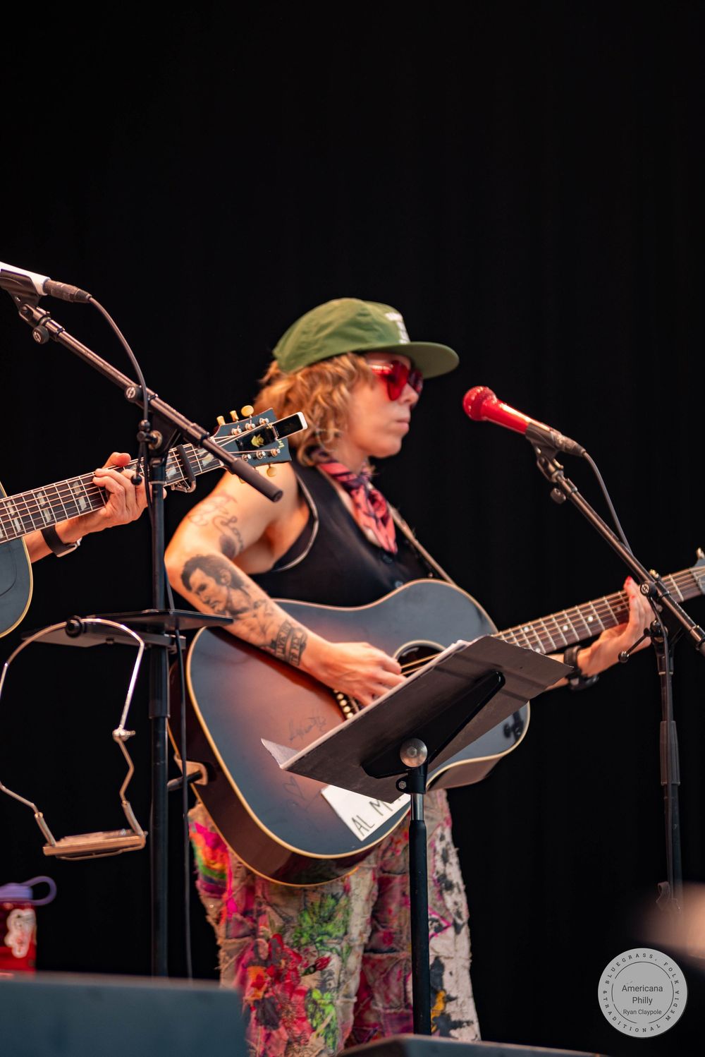 Tattooed female guitarist in green cap and red sunglasses performs acoustic guitar on stage at outdoor music festival.