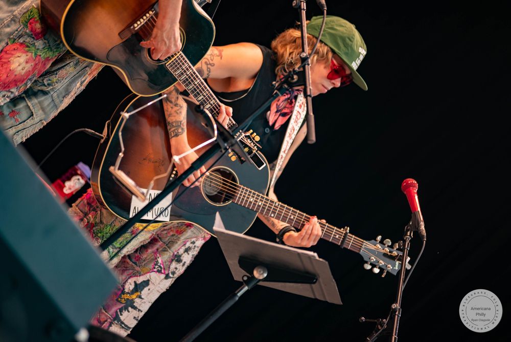 Two tattooed musicians playing acoustic guitars on stage under warm lighting at a live concert.