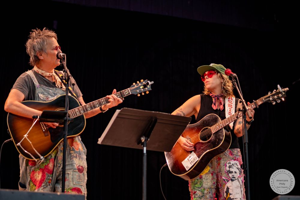 Two women playing acoustic guitars and singing on stage at an outdoor music festival performance.