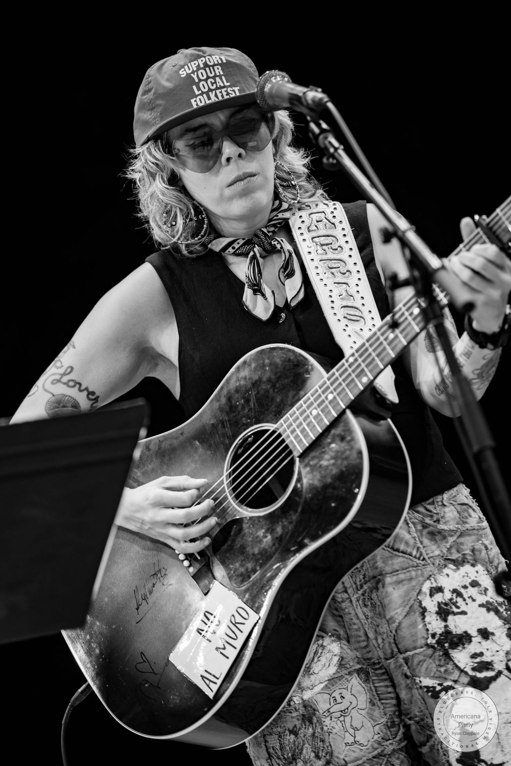 Black and white photo of a musician playing acoustic guitar at a microphone, wearing a cap and bandana on stage.