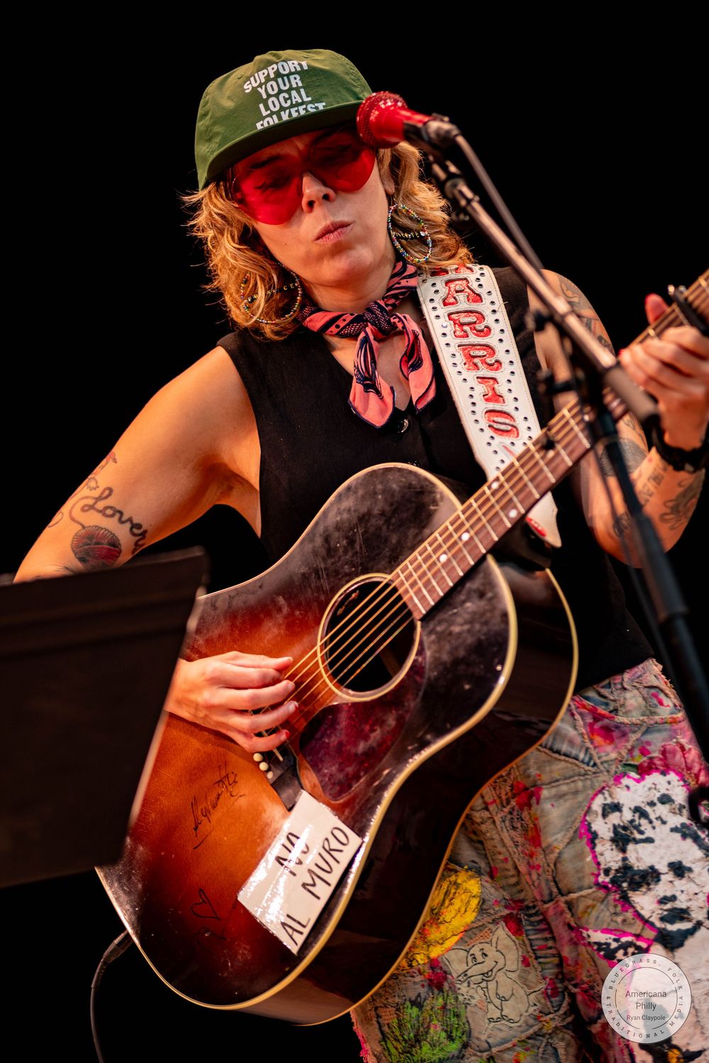 Female musician playing acoustic guitar on stage, wearing green cap, red sunglasses, and floral pants at a live performance.