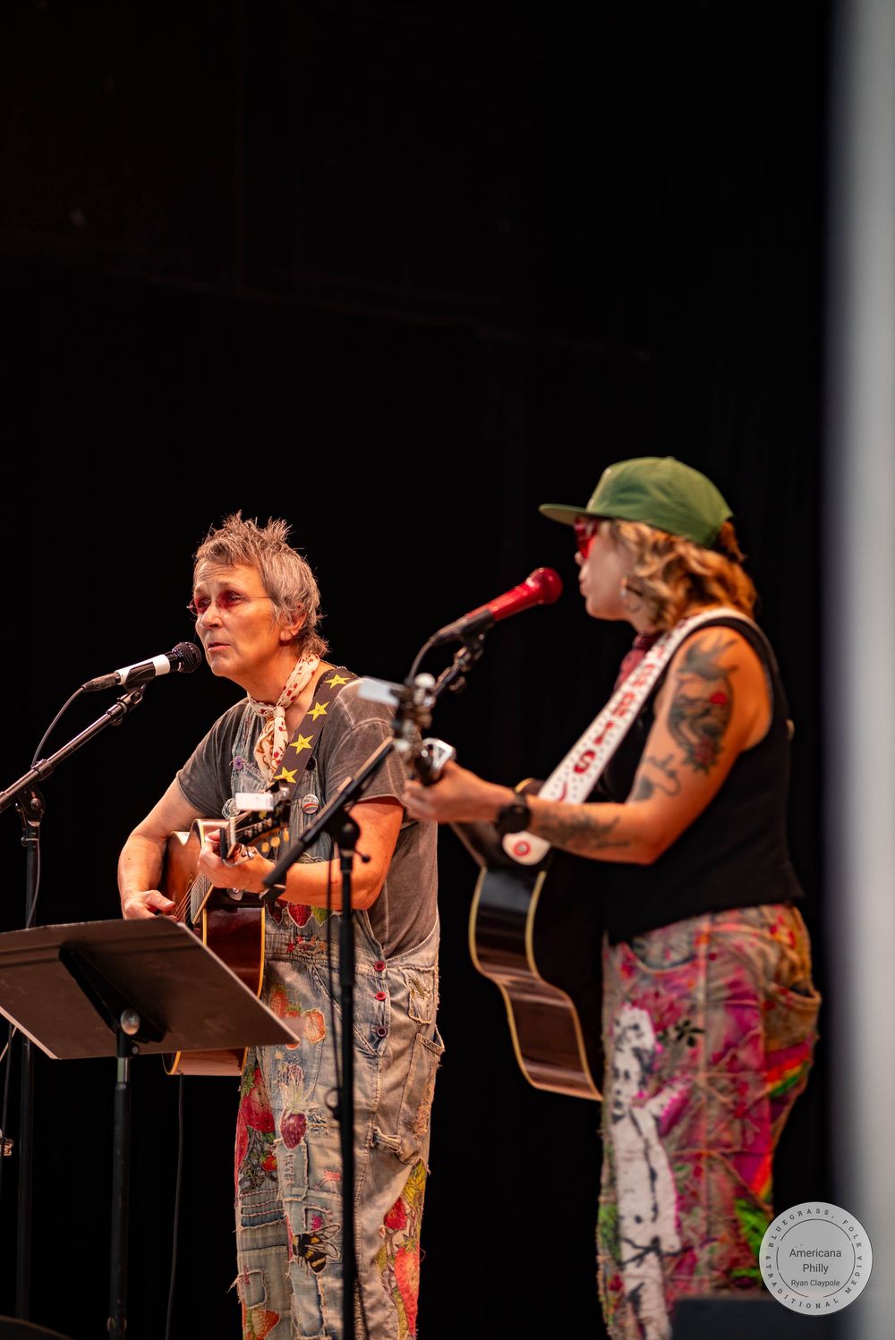 Two women playing acoustic guitars and singing into microphones on stage, one with tattoos wearing a green cap and colorful pants.