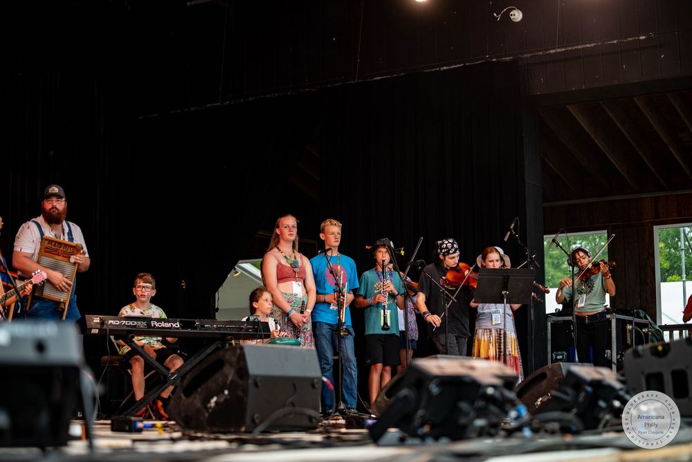 A folk band performs on an outdoor stage, featuring violin, keyboard, and various instruments at a summer music festival.