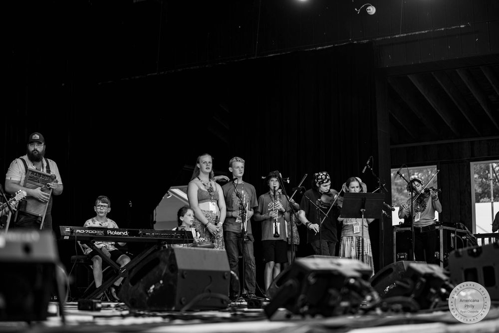 Black and white photo of a folk band performing on an outdoor stage with multiple musicians playing instruments.