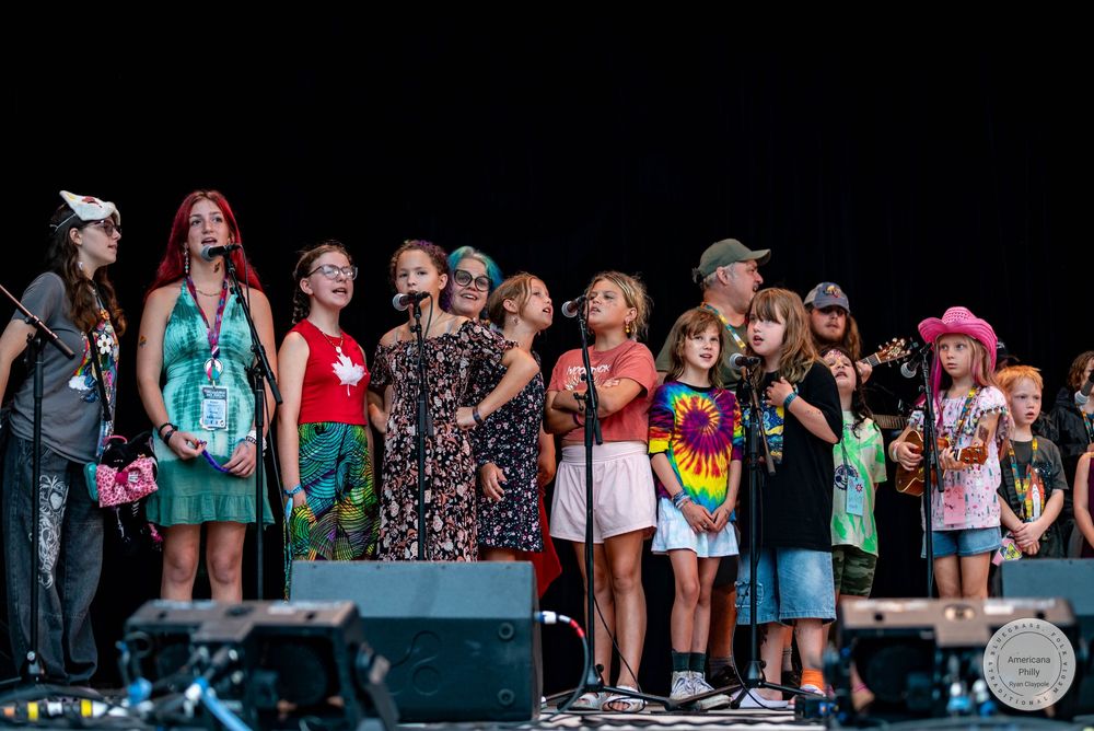 Group of adults and children performing on an outdoor stage at a summer music festival.
