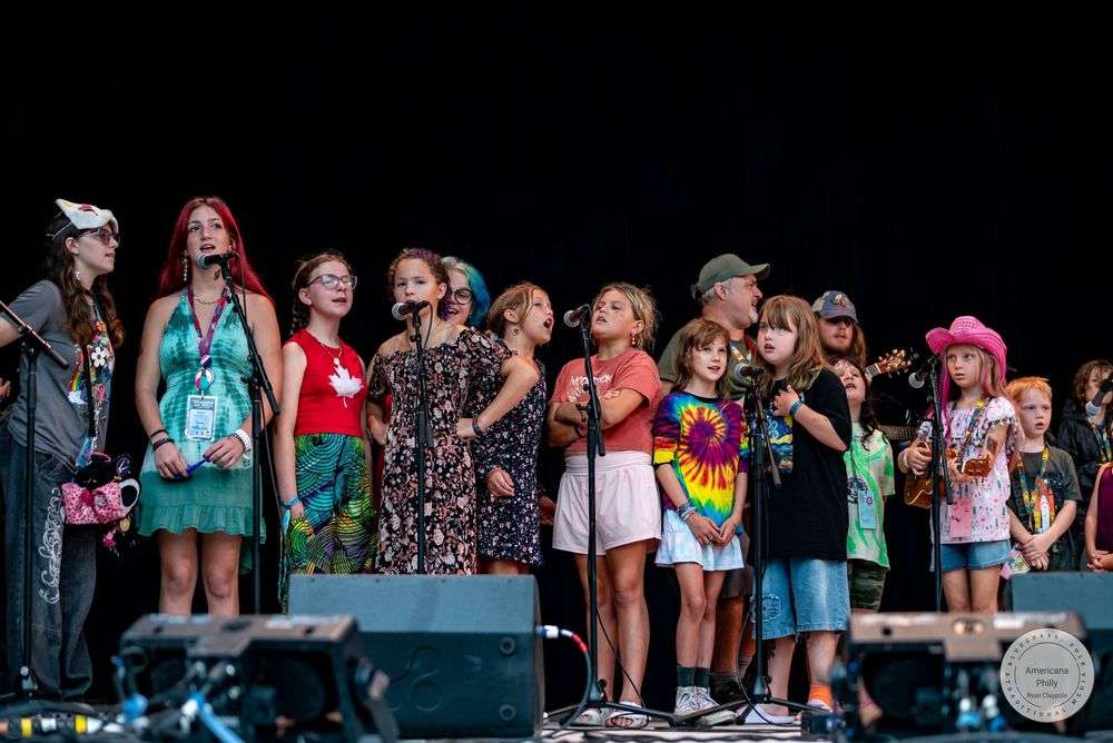 Children and adults perform together on an outdoor festival stage, singing into microphones under bright summer skies.